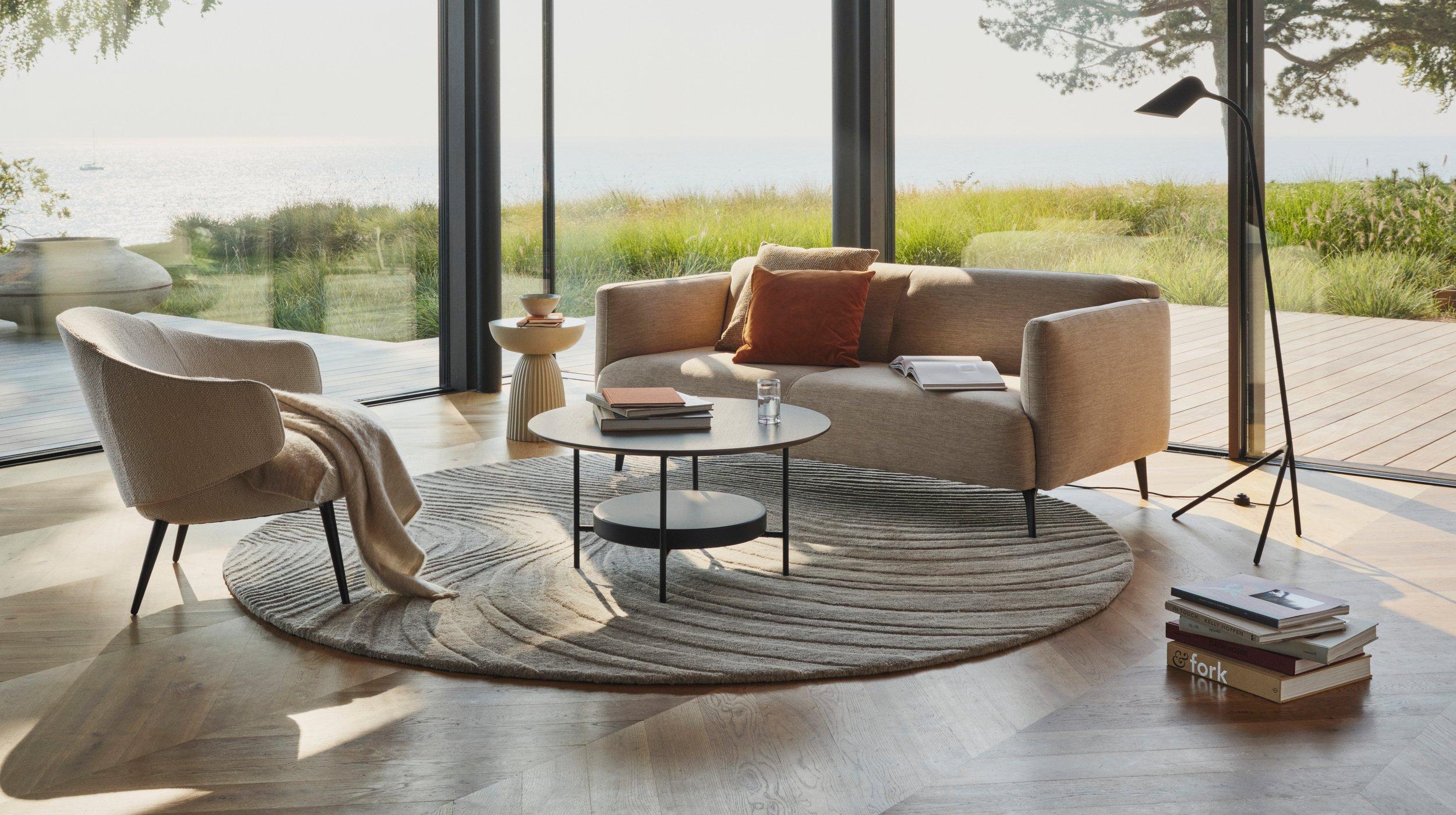 现代起居室配有米色 Modena 沙发，摆在条纹地毯上的圆形咖啡桌，还有可以欣赏户外葱郁景观的落地窗。